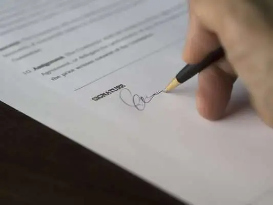 A hand holding a pen signing a document with a visible signature line.