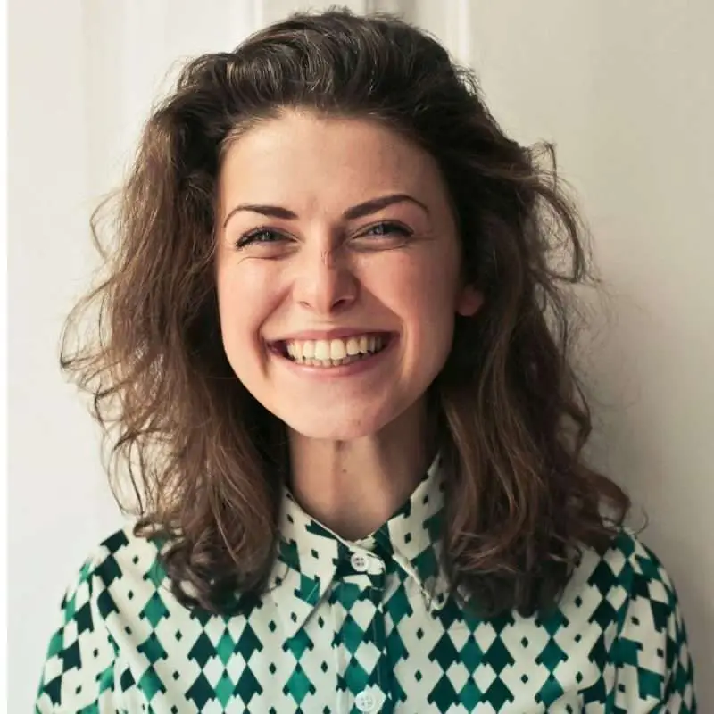 A smiling woman with curly hair wears a patterned shirt and looks directly at the camera against a light wall.