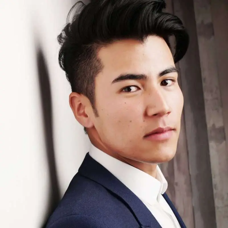 A young man in a navy suit and white shirt faces the camera with a calm expression and neatly styled dark hair.